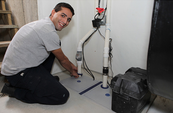 Man in a gray shirt smiling while installing a basement sump pump system. He is kneeling beside white PVC pipes and a black plastic container.