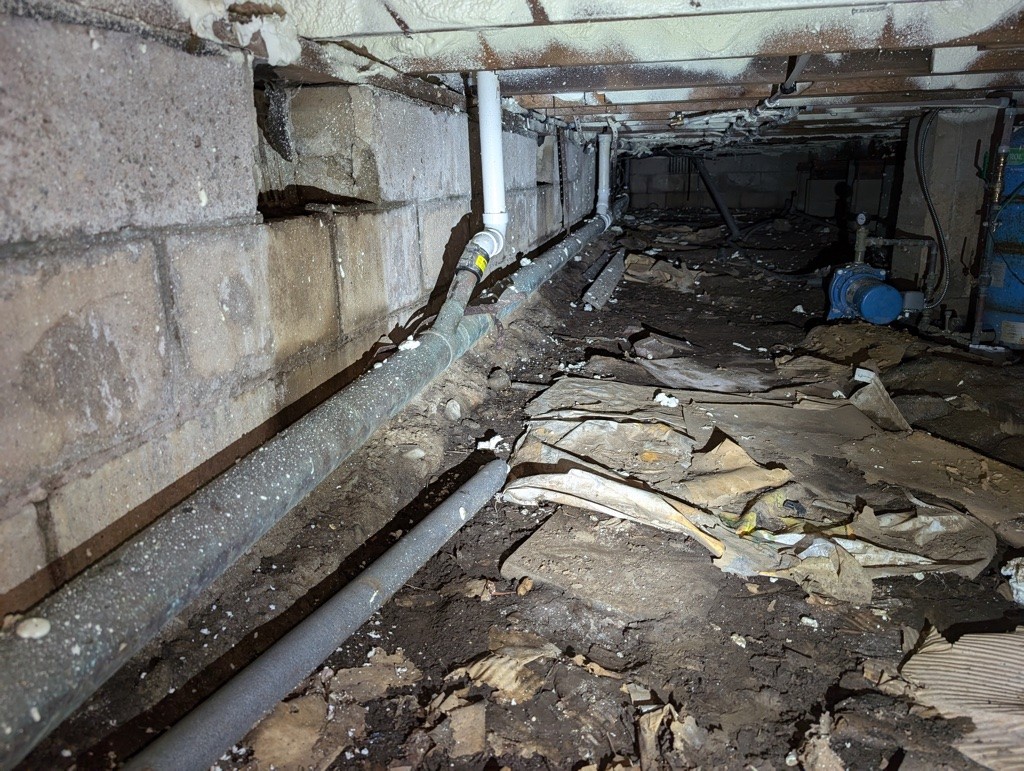 Dimly lit crawl space with exposed pipes along a concrete wall. The ground is cluttered with debris and dirt, creating an abandoned, neglected feel.